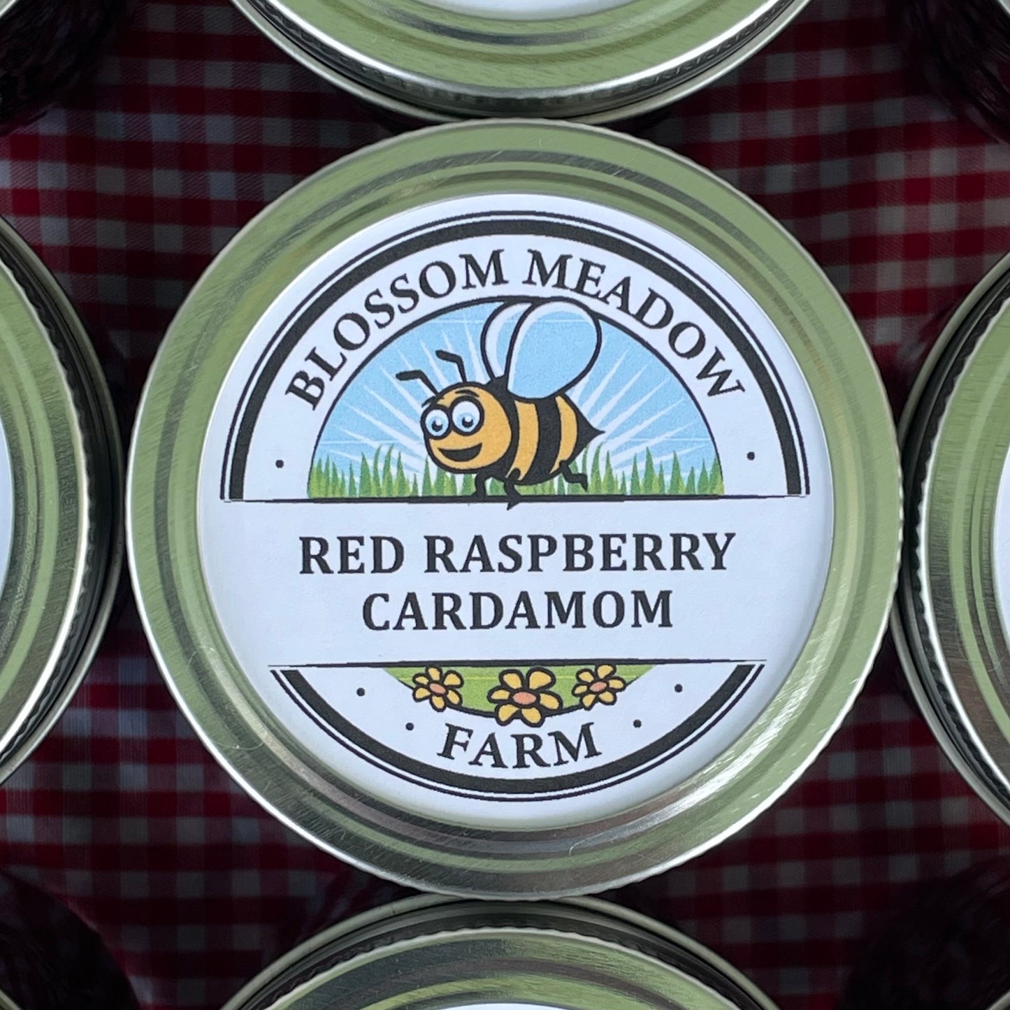 A close-up of a round metal can lid with a printed label reading 'Blossom Meadow Red Raspberry Cardamom Farm' and a cartoon bee on a red gingham background.