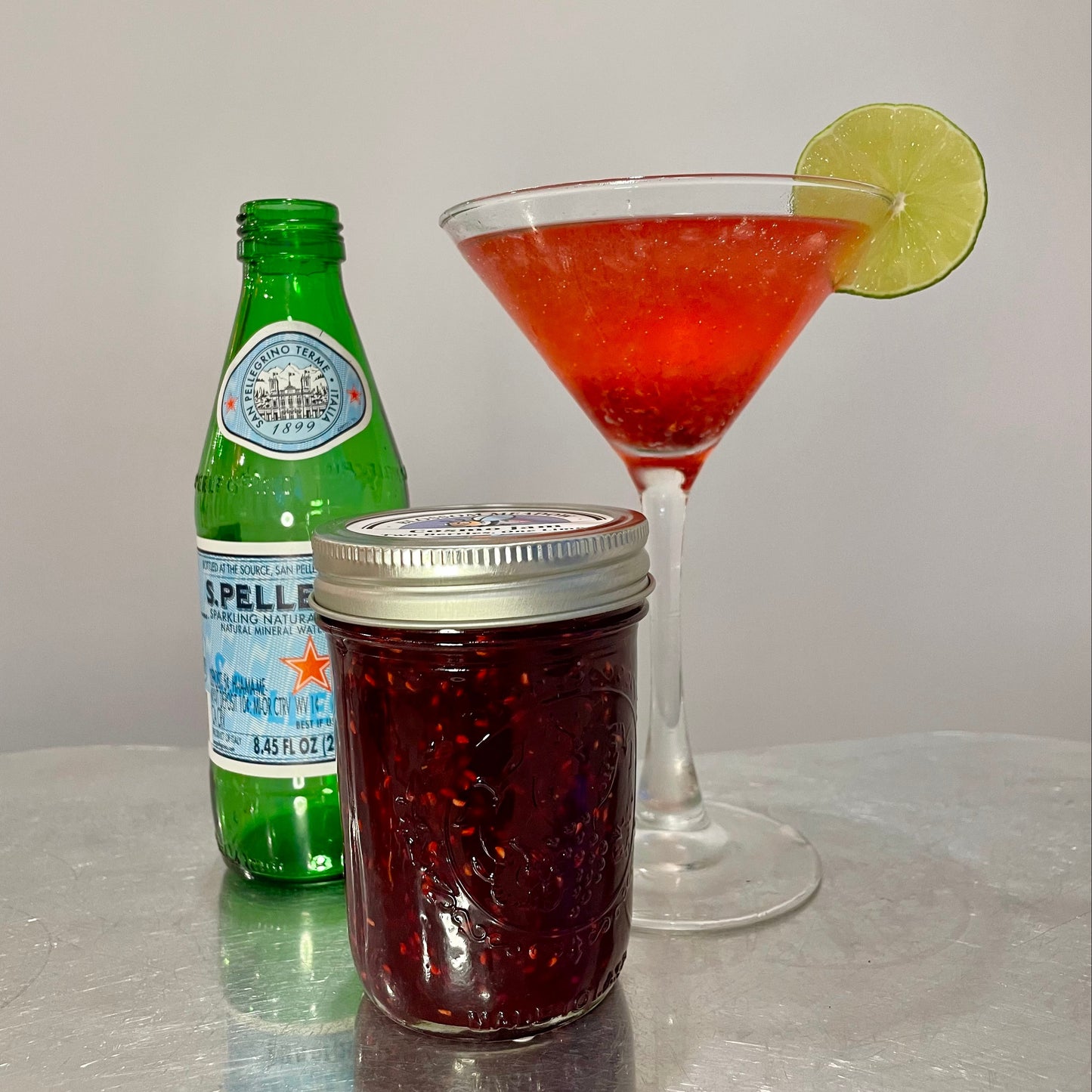 Cosmo Jam Gazoz Mocktail in a martini glass with a bottle of San Pellegrino and a jar of Blossom Meadow Farm Cosmo Jam on a reflective surface.