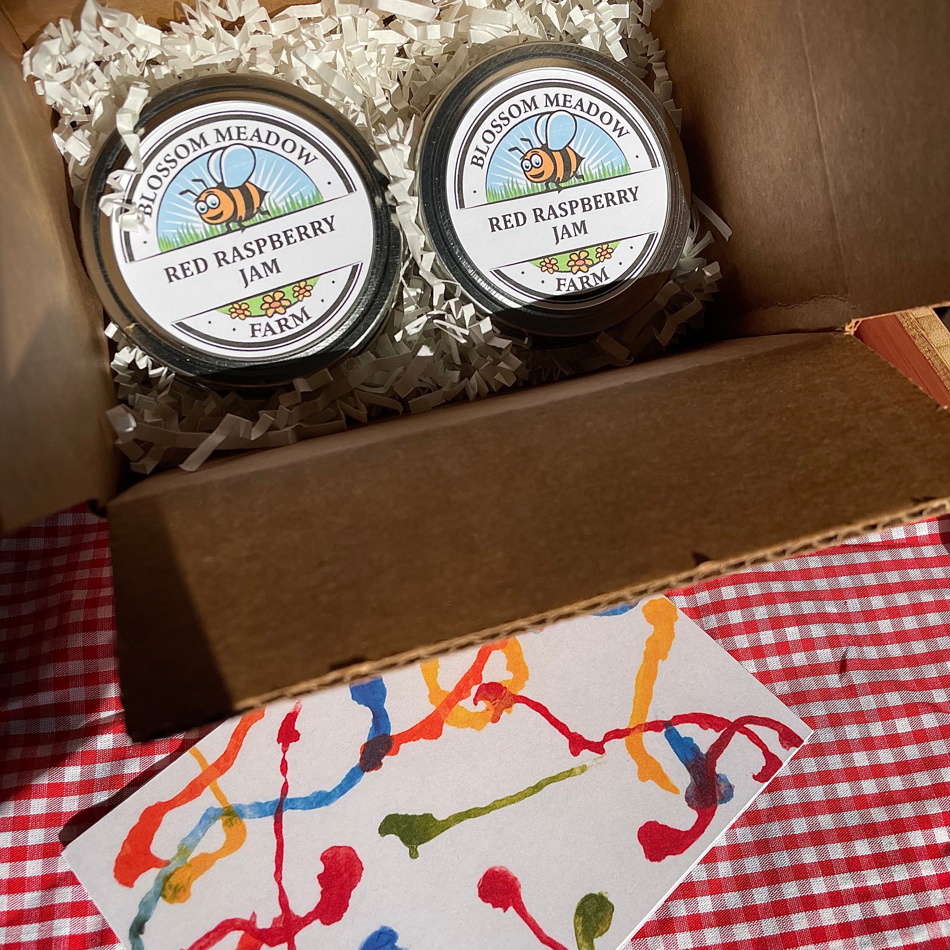 Two jars of Blossom Meadow Red Raspberry Jam in a box on a red and white checkered tablecloth.
